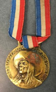 A bronze colored medal depicting the head and shoulders of Jacob Leisler wearing a brad-brimmed hat. A red, white, and blue ribbon is attached at top.