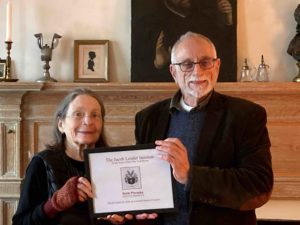 Ruth Piwonka and David Voorhees stand in front of a fireplace mantel holding a certificate recognizing Ruth's contributions to teh Insitute.